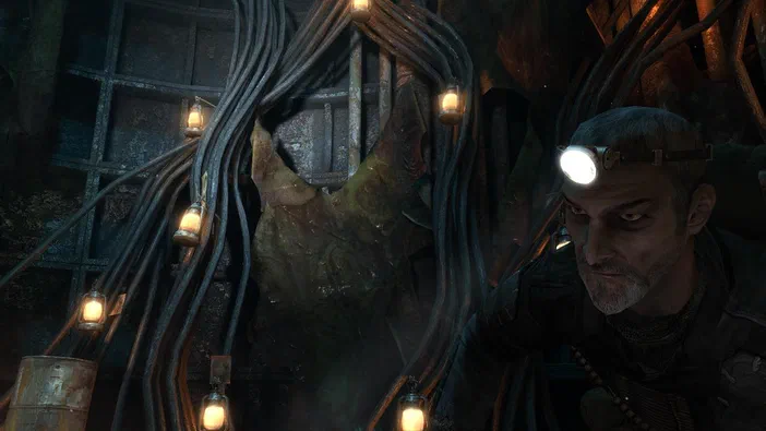 Dans le jeu vidéo Metro 2033, un homme portant une lampe frontale se tient devant des câbles suspendus avec des ampoules dans un décor post-apocalyptique sombre.