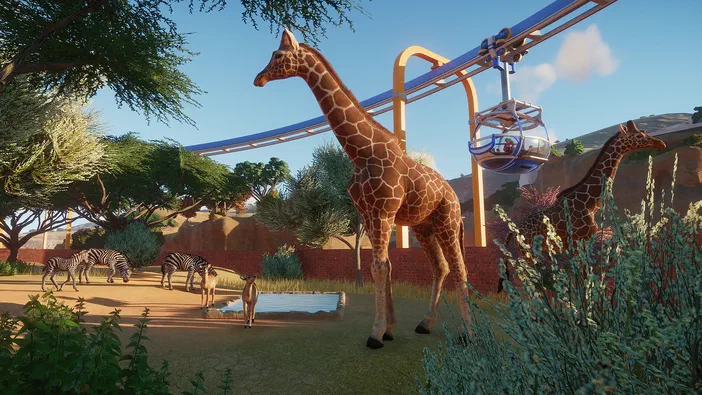 Capture d'écran du jeu Planet Zoo montrant des girafes et des zèbres dans un enclos avec une montagne en arrière-plan.