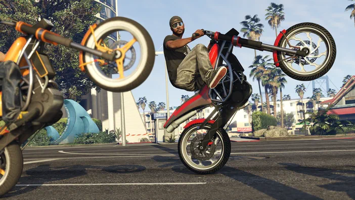 Dans Grand Theft Auto V Legacy, un personnage effectue une roue de moto sur une rue ensoleillée avec des palmiers et d'autres véhicules à l'arrière-plan.